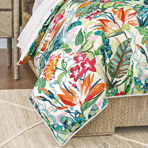 A tropical floral print comforter draped over a bed frame in a styled bedroom setting, featuring orange, pink, green, and blue botanical motifs on a cream background.